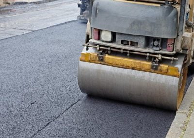 A road roller compacts fresh asphalt on a newly paved road, with traffic cones and workers visible in the background, showcasing expert asphalt paving techniques.