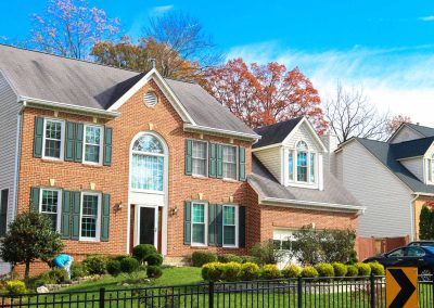 Two-story brick house with large windows and a front yard featuring manicured bushes and asphalt paving, set against the backdrop of autumn trees in Virginia.