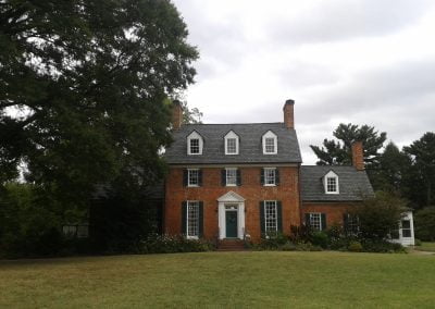 A large brick colonial house with multiple chimneys, dormer windows, and a green lawn under a cloudy Manassas, VA sky.