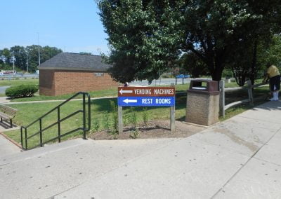 A signboard at a Virginia rest stop points left to vending machines and right to rest rooms, with trees and freshly laid asphalt paving in the background.