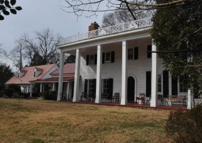 A large white colonial-style house with a red roof, tall columns, a porch, and a lawn surrounded by trees, located in the charming town of Manassas, Virginia. The property features elegant paving leading up to the grand entrance.