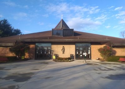 A church with a triangular roof and cross, brick exterior, and glass double doors is surrounded by trees and a paved entrance. Nestled in the heart of Manassas, Virginia, the serene setting features well-maintained asphalt paving.