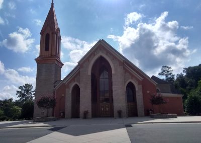 A church with a tall steeple, pointed arches, and a cross on top stands under a partly cloudy sky above the asphalt paving in Manassas, VA.