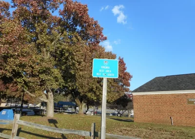 A green sign for a Virginia Rest Area, Mile 155 South, stands next to trees and a brick building under a clear blue sky, with the well-maintained asphalt adding a touch of modernity to the scenic view.