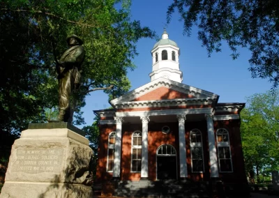 A historic red-brick courthouse with a white bell tower and a Confederate soldier statue in front, standing proudly under the blue sky in Manassas, VA, with an asphalt pathway leading up to it.